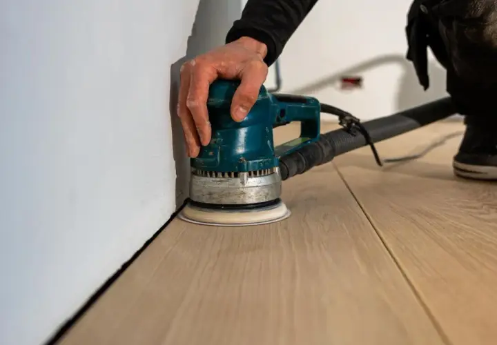 Mano che usa una levigatrice orbitale per levigare il parquet fai da te, avvicinandosi al battiscopa bianco.