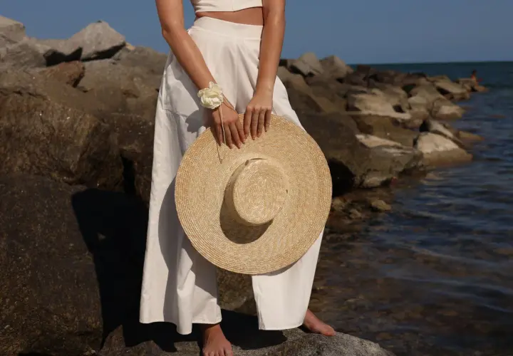 Mujer con un sombrero para ir a los toros, vestida de blanco, de pie sobre rocas junto al mar.