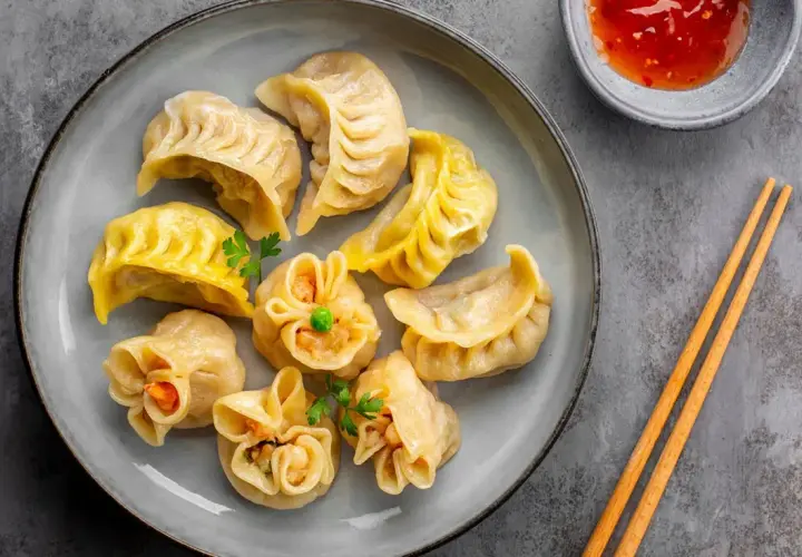 Talerz pełen różnorodnych pierożków dim sum, z sosem i pałeczkami.