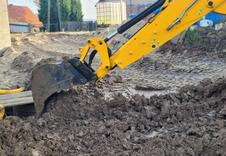 Żółta koparka marki JCB pracuje na budowie, kopiąc ziemię. Widać jej ramię i łyżkę zagłębiającą się w błotniste podłoże.