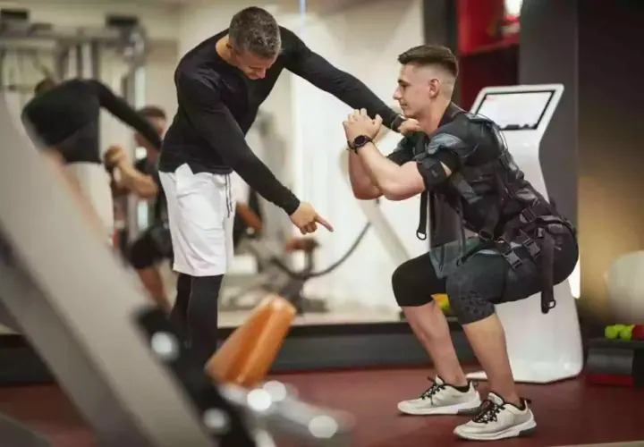Trener instruuje mężczyznę w kamizelce EMS podczas ćwiczeń w siłowni.