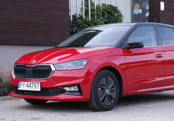 Czerwona Skoda Fabia 4 z czarnym dachem i felgami stoi przed budynkiem z drewnianą elewacją.