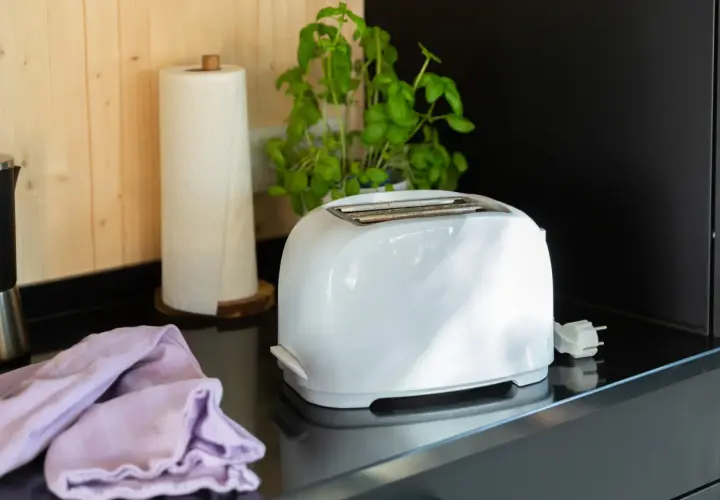 Ein weißer Toaster steht auf einer schwarzen Arbeitsplatte, bereit für die Reinigung. Daneben liegen ein lila Tuch und eine Küchenrolle.