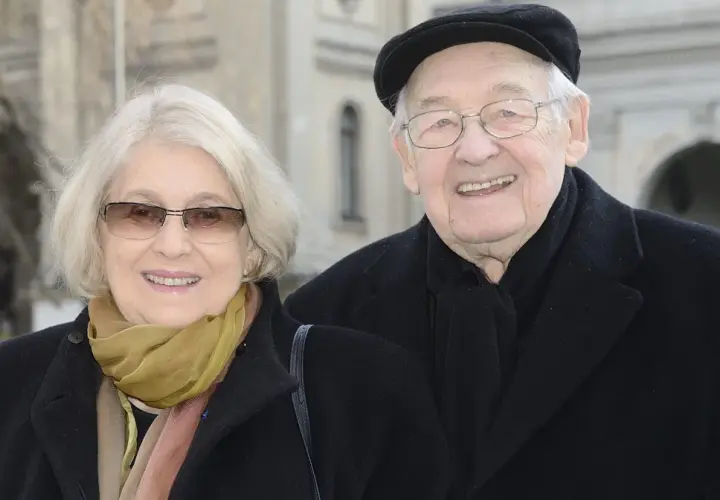 Uśmiechnięta para: Andrzej Wajda i jego żona, w eleganckich płaszczach i szalikach, pozują do zdjęcia na tle architektury.