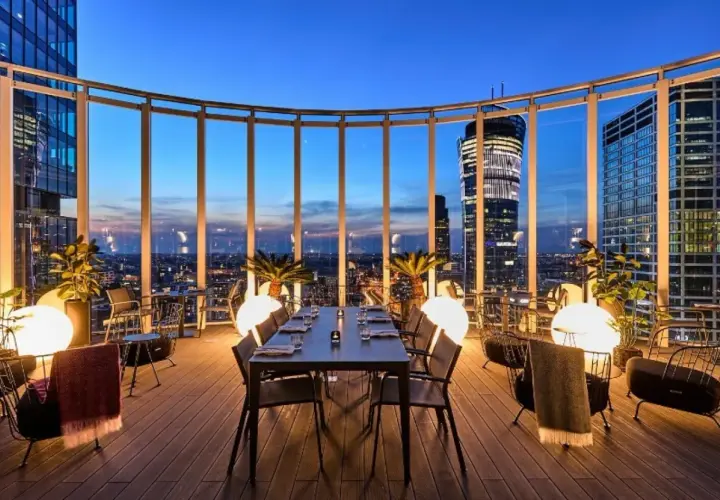 Przytulna restauracja Warszawa na dachu z widokiem na nocne miasto. Stoły i krzesła, palmy i kule świetlne tworzą magiczną atmosferę.