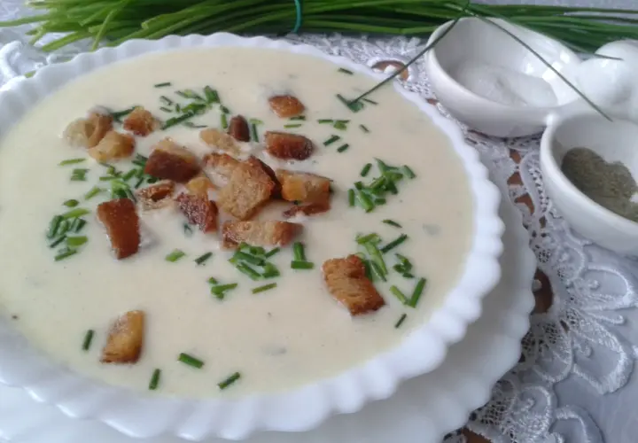 Kremowa zupa porowa z grzankami i szczypiorkiem, podana w białej miseczce. Obok małe miseczki z solą i pieprzem.