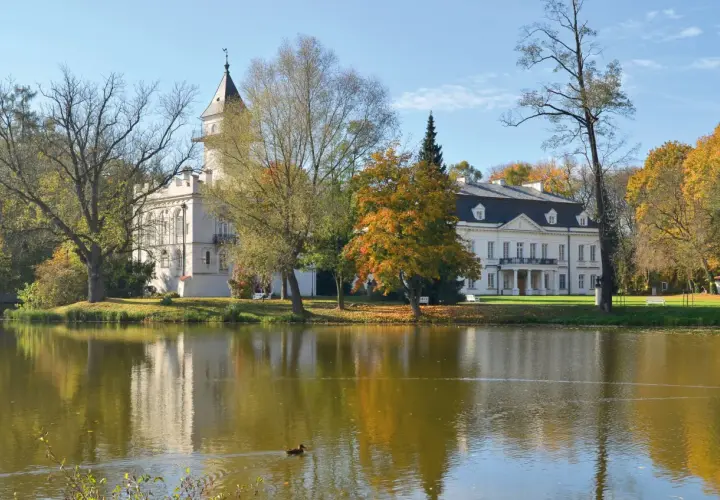 Pałac w jesiennej scenerii, otoczony drzewami o barwnych liściach, odbija się w spokojnej tafli jeziora. W oddali widać fragment puszczy bolimowskiej.