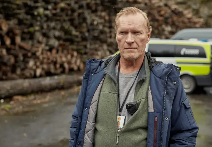En man med intensiv blick, klädd i jacka och väst, bär en radio och ett id-kort. Bakom honom syns en polisbil och en vedhög. En scen från en norsk serie.