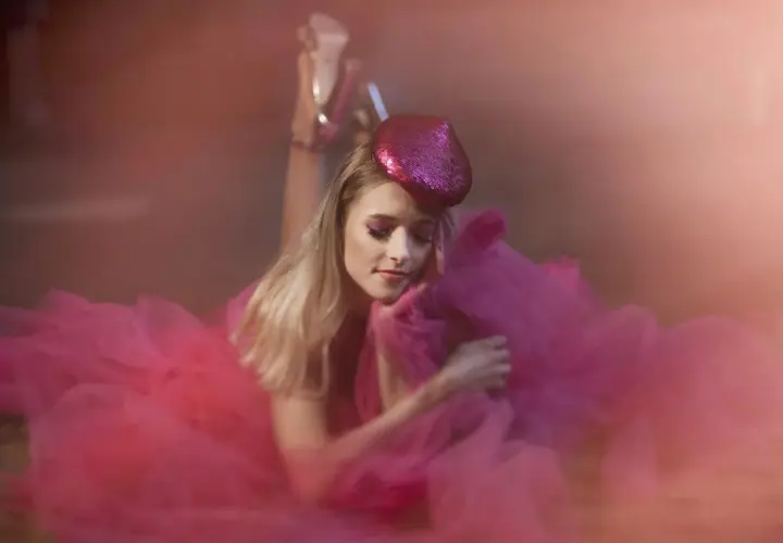 Urocza sesja glamour: blondynka w różowej tiulowej sukni i błyszczącej beretce, z makijażem oczu w tym samym kolorze, leży na ziemi.