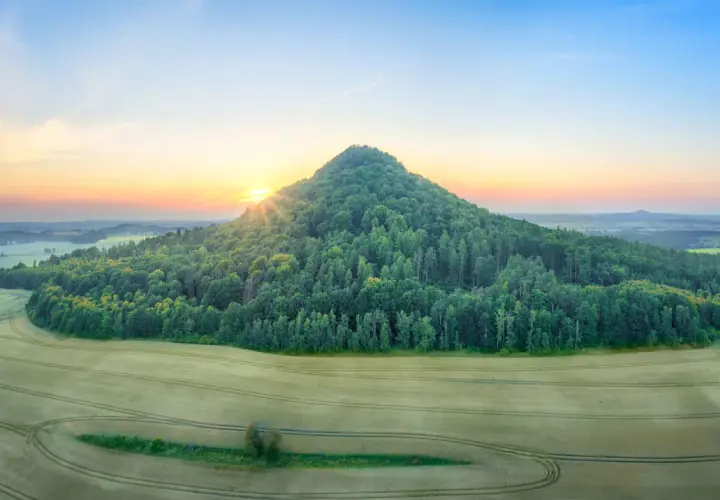 Zalesione wzgórze przypomina wygasłe wulkany w Polsce. Słońce wschodzi nad krajobrazem pól i lasów.