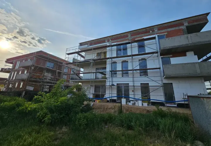 Budowa nowych bloków mieszkalnych, widać rusztowania i izolację. Słońce przebija się przez chmury.