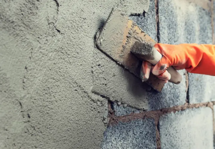 Mano con guanto arancione stende malta per intonaco esterno su un muro di blocchi di cemento con una cazzuola.