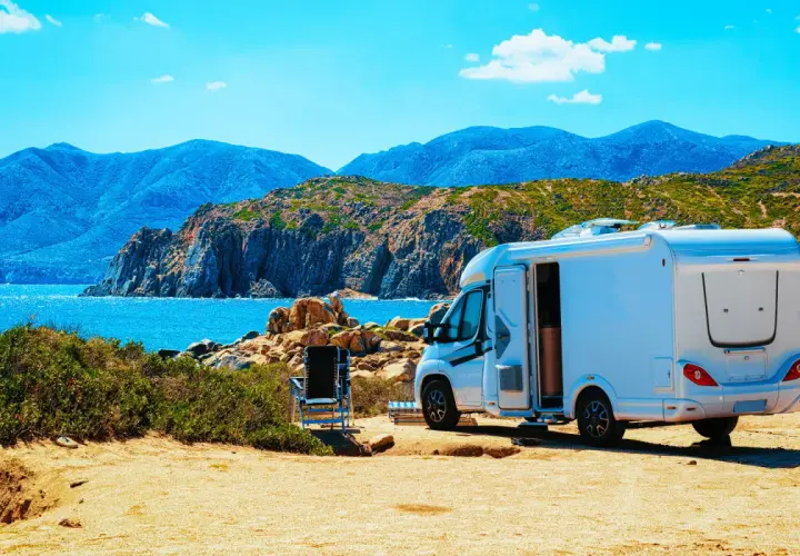 Un camping-car blanc est garé sur un terrain sablonneux, face à une mer bleue et des montagnes. L'application spot camping car idéale pour ce paysage.