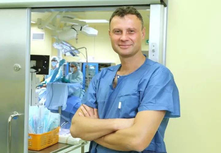 Lekarz w niebieskiej koszuli chirurgicznej stoi na tle sali operacyjnej.