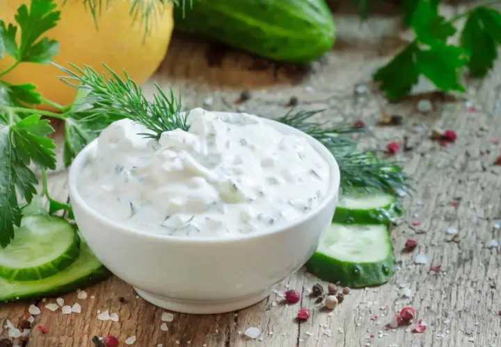Pyszny dip jogurtowy z ziołami i ogórkiem, udekorowany koperkiem, podany z plastrami ogórka i przyprawami na drewnianym blacie.