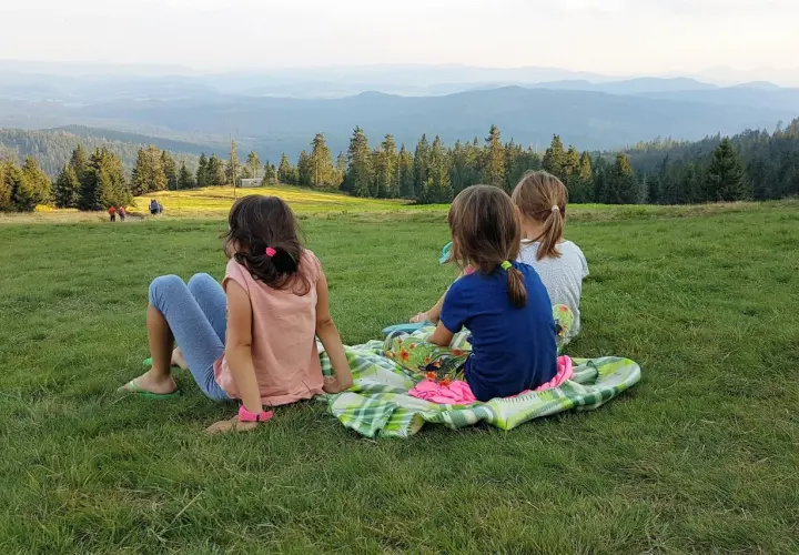 Trzy dziewczynki, jak małe rysianki, siedzą na trawie, podziwiając widok gór. Jedna z nich, z żabnicą na głowie, wpatruje się w dal.