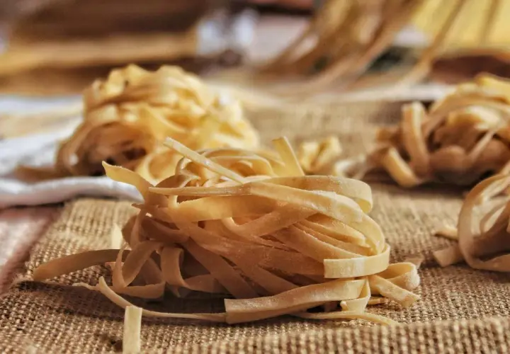 Nidi di tagliatelle fatte in casa, pronte per essere cucinate. La pasta fresca, con la sua texture rustica, è disposta su una tela grezza, invitando a un pasto autentico.