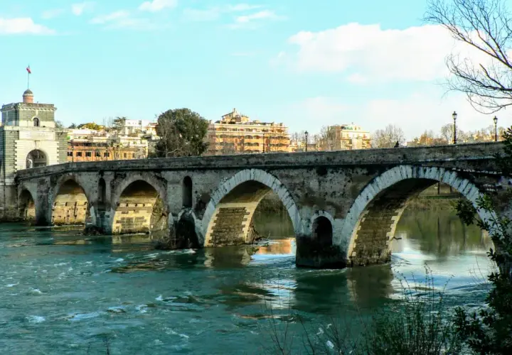 Il **puente Milvio** attraversa il Tevere con i suoi archi antichi, mentre sullo sfondo si stagliano palazzi romani sotto un cielo azzurro.