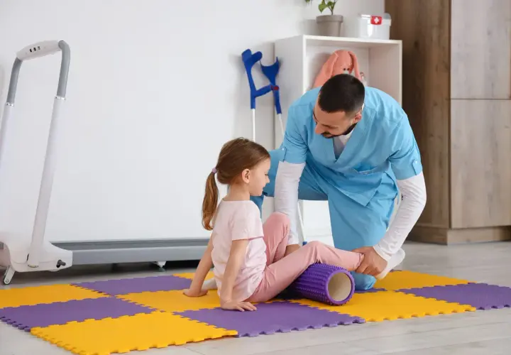 Fizjoterapeuta stosuje metodę Bobath podczas ćwiczeń z dziewczynką, która leży na macie z wałkiem pod nogą.