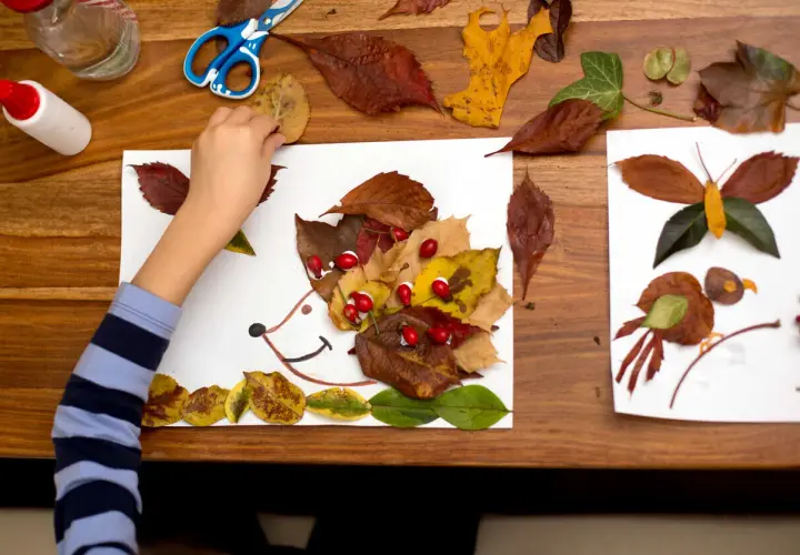 Niño haciendo otoño manualidades preescolar: un erizo con hojas secas y bayas rojas, junto a una mariposa y un pájaro hechos de hojas.