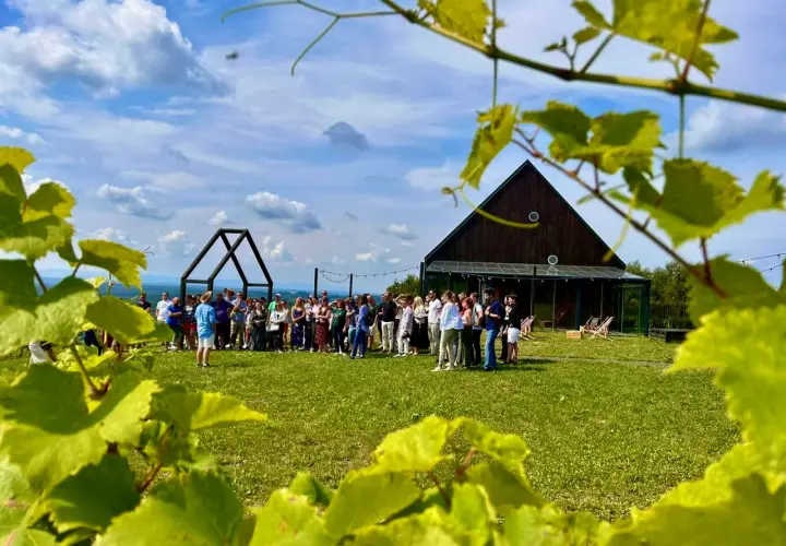 Grupa ludzi zwiedza winnicę, poznając tajniki produkcji wina. W tle nowoczesny budynek i malowniczy krajobraz.