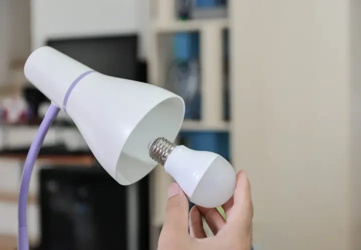 Ręka wymienia żarówkę w lampce biurkowej, symbolizując modernizację oświetlenia i oszczędność energii.