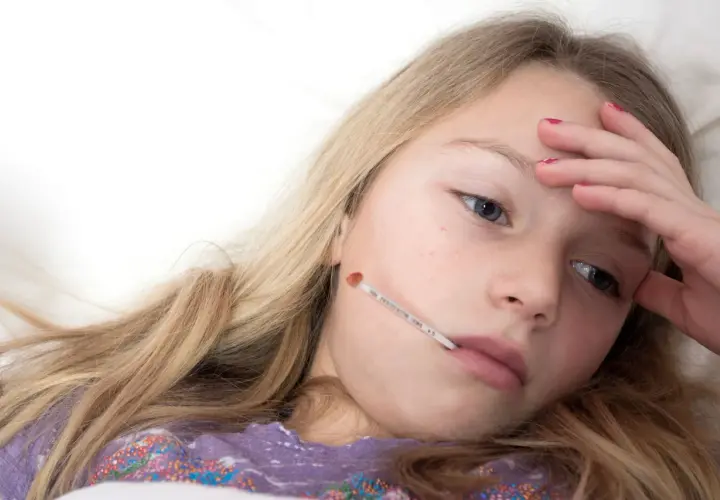 Une jeune fille malade avec un thermomètre dans la bouche, se demandant combien de temps dure la mononucléose.