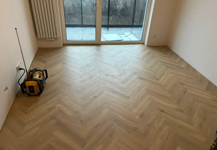 Nowy, drewniany panel winylowy ułożony we wzór jodełki. W rogu stoi żółto-czarne urządzenie.