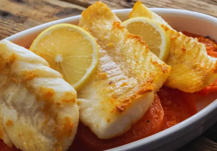 Filetes de pescado blanco horneado con rodajas de limón y tomates en una fuente blanca.