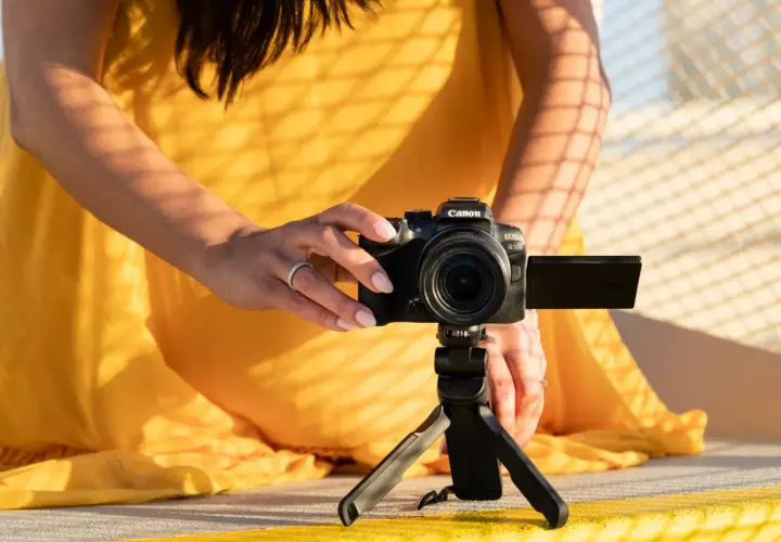 Kobieta w żółtej sukience ustawia aparat Canon EOS R10 na statywie, eksperymentując z trybem manualnym w aparacie.