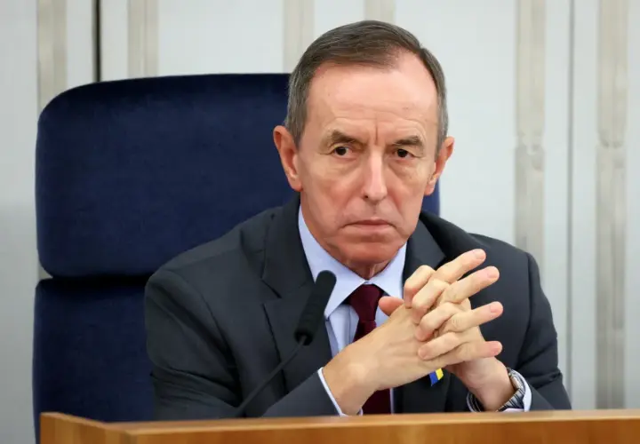 Tomasz Grodzki w garniturze, z rękami złożonymi na piersi, siedzi przy stole. Na klapie marynarki przypinka Poczty Polskiej.