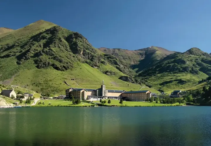 Lago sereno frente a un complejo de edificios en Queralbs, rodeado de montañas verdes y un cielo azul claro.