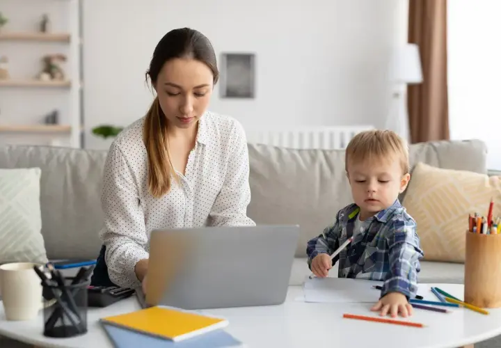 Kobieta pracuje na laptopie, podczas gdy jej mały syn rysuje obok. To codzienna opieka na dziecko i praca zdalna w jednym.
