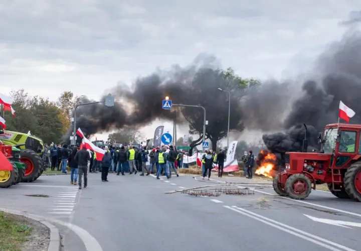 Czerwony traktor na drodze, w tle dym i ludzie z polskimi flagami.