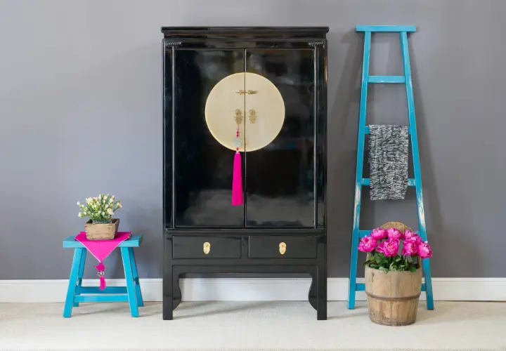 Mobile con laccatura nera lucida, dettagli dorati e nappa fucsia. Accanto, sgabello blu con fiori e scaletta decorativa.