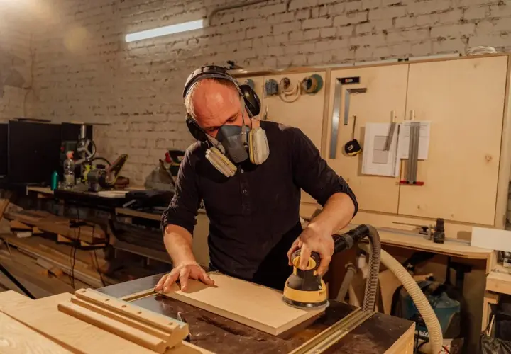 Artisan concentré, masqué et casqué, réalisant un ponçage bois méticuleux avec une ponceuse électrique dans son atelier.