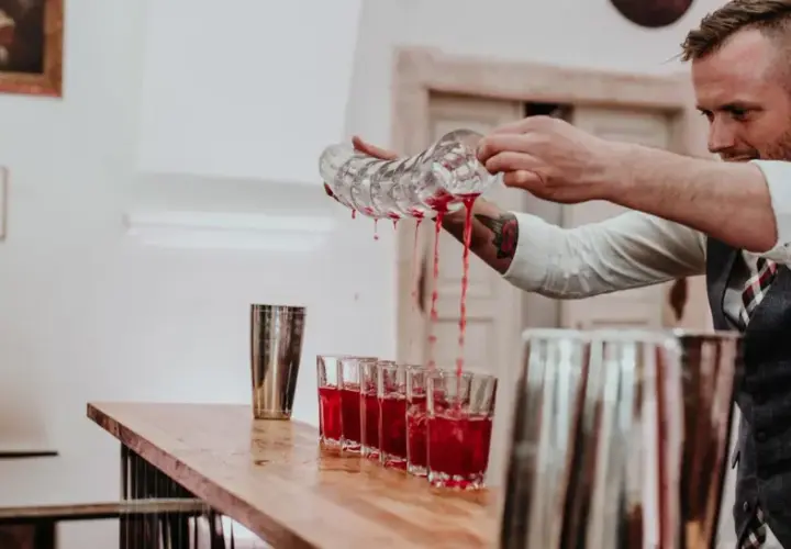 Barman nalewa czerwony napój do kieliszków na weselu, obok podekscytowana panna młoda.