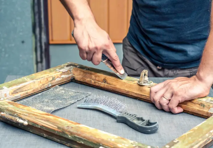 Mani esperte usano uno raschietto per sverniciare legno, rimuovendo vecchie vernici da un telaio. Accanto, una spazzola metallica e carta vetrata.