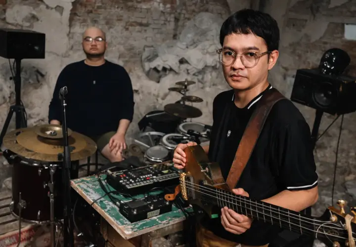 Dwóch muzyków w surowym wnętrzu, jeden gra na gitarze, drugi siedzi przy perkusji. Brzmienie tutti jest gotowe do nagrania.