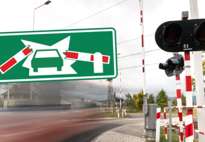 Zielony znak informuje o oznaczeniu przejazdu kolejowego. Po bokach widać rogatki i sygnalizację świetlną.