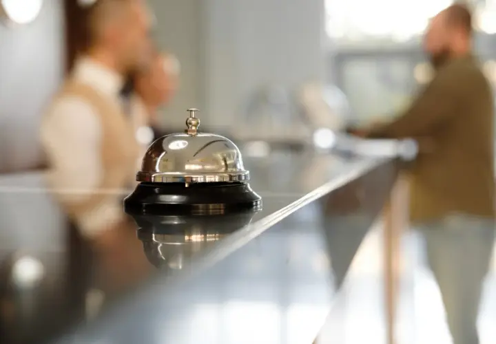 Dzwonek hotelowy na ladzie, symbol usług uzupełniających. W tle niewyraźne postacie personelu.