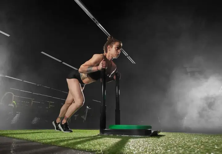 Kobieta z tatuażami pcha sanie treningowe. To właśnie crossfit co to – siła, wytrzymałość i determinacja w akcji.