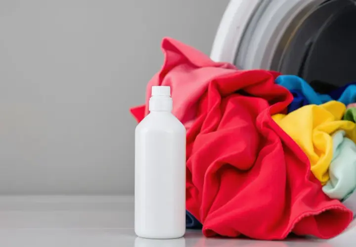 Bouteille de vinaigre blanc à côté d'une machine à laver remplie de linge coloré. Idéal pour enlever la colle tenace des textiles.