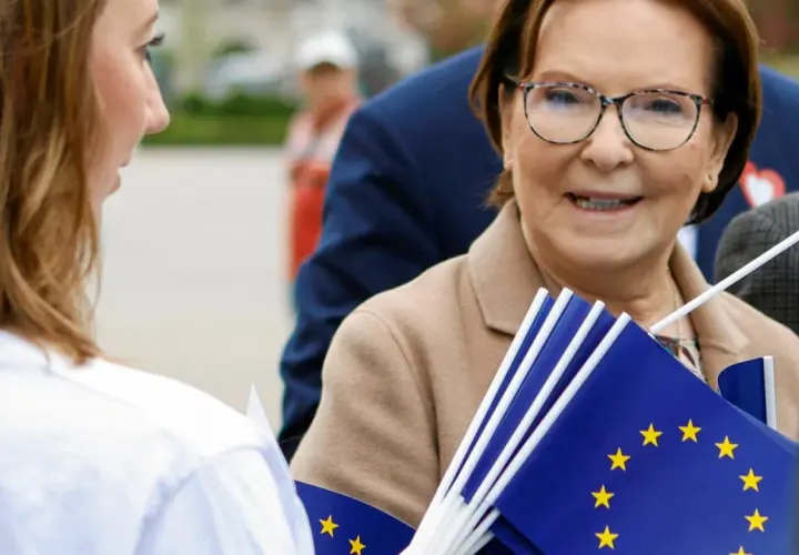 Ewa Kopacz, gdzie mieszka, uśmiecha się, trzymając flagi UE.
