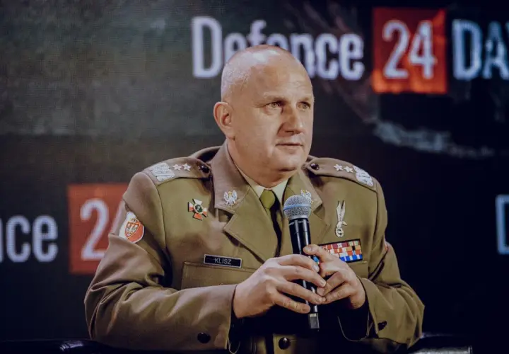 Generał Klisz w mundurze, z mikrofonem w dłoniach, siedzi przed ekranem z napisem "Defence 24 DAYS".