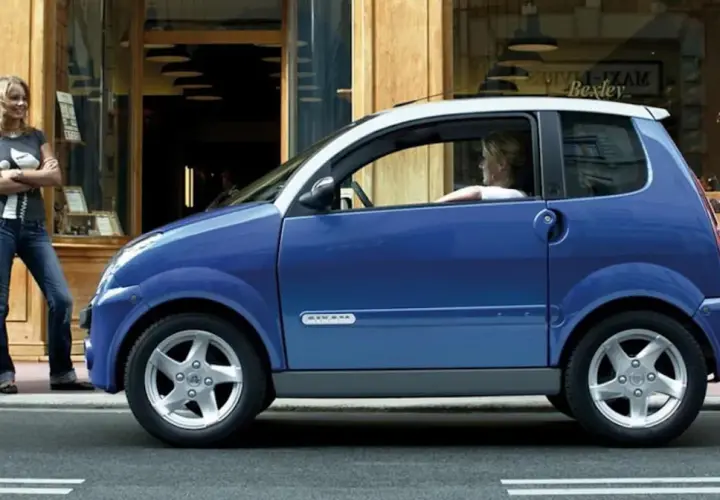Niebieskie auto od 14 lat, z kobietą za kierownicą, stoi przed sklepem. Obok niego stoi druga kobieta.