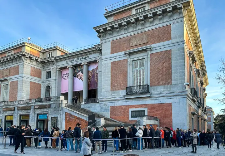 Kolejka turystów przed wejściem do Muzeum Prado w Madrycie. Monumentalna fasada budynku z cegły i kamienia ozdobiona jest banerami wystawowymi.