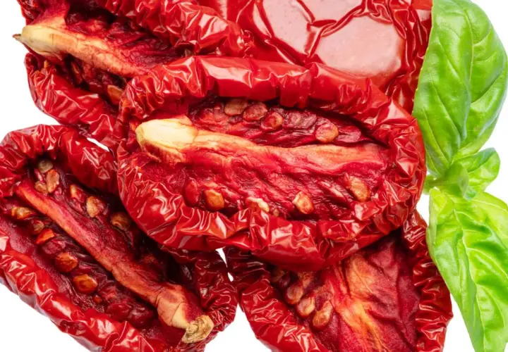 Tomates secos al horno, arrugados y rojos, con semillas visibles, junto a una hoja de albahaca verde fresca.