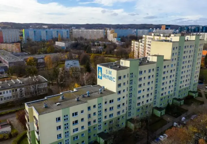 Duży blok mieszkalny S.M. Młyniec z charakterystycznym logo, otoczony innymi budynkami i drzewami jesienią.