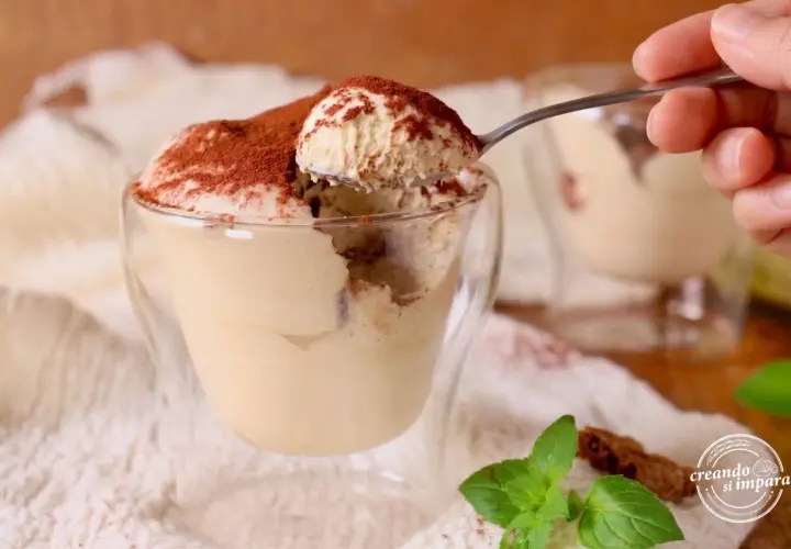 Un cucchiaino preleva una porzione generosa di soffice mousse al caffè, cosparsa di cacao, da un bicchiere di vetro.